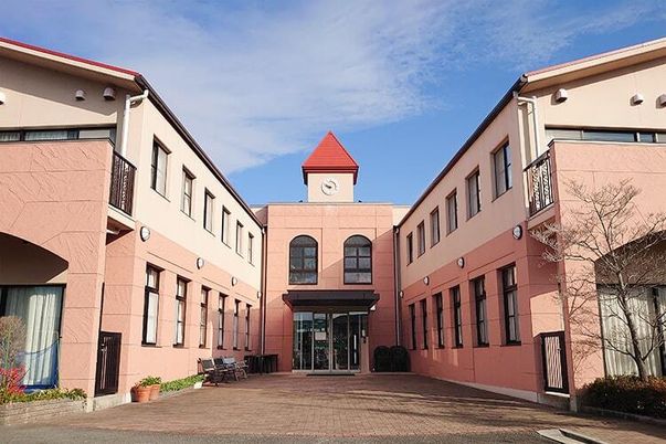 ピンクのコの字型の２階建ての建物は日当たり良好で建物の周りには草木が植えられ、プランターも多く置かれている。