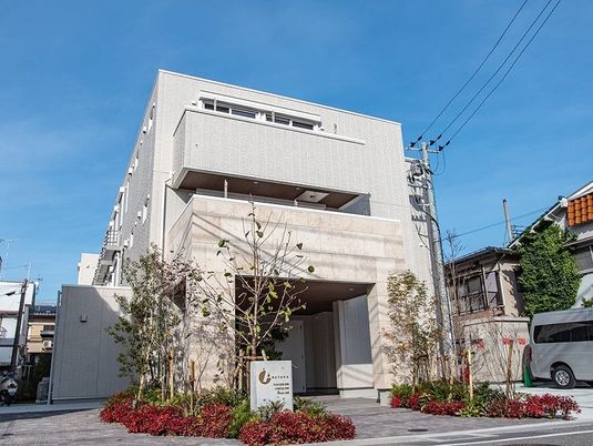 ライトグレーの3階建ての建物の周りには赤く染まった低木やほっそりとした木々が植えられ、駐車スペースもある。