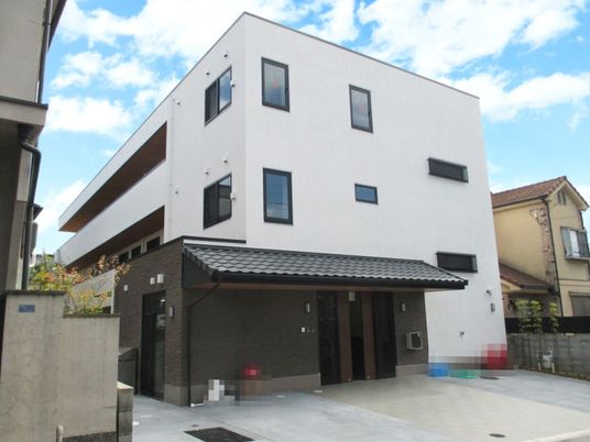 白い3階建ての建物。2階と3階にはベランダが設置され、玄関には瓦屋根が付き、駐車場もある。