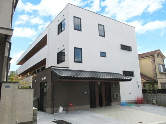 白い3階建ての建物。2階と3階にはベランダが設置され、玄関には瓦屋根が付き、駐車場もある。