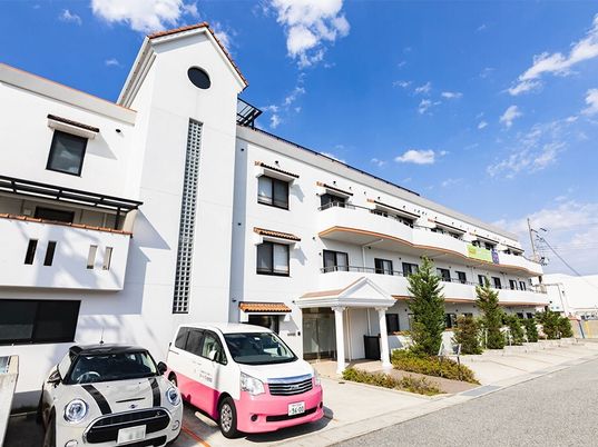 白を基調とした3階建ての建物の前には駐車スペースがあり、草木も植えられている。