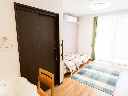 寝具付きのベッドが置かれたフローリングの部屋には、ラグが敷かれ、エアコンや車いす対応の洗面所もある。