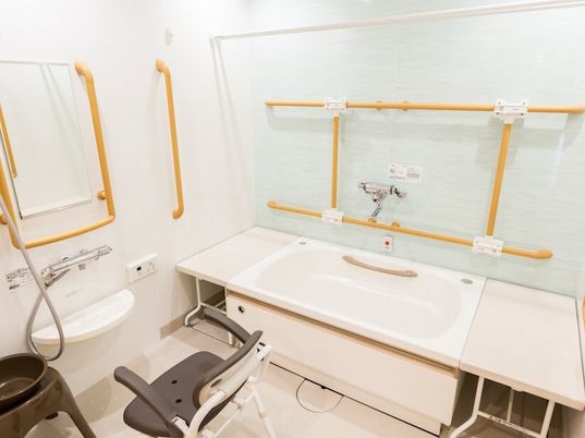 手すりがたくさん付いた浴室には一人用の浴槽が設置され、シャワーもあり、オレンジの呼び出しボタンやベンチもある。