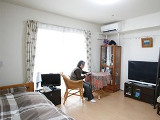 テレビやベッド、エアコンなどの家具家電がそろった居室でくつろいで過ごす入居者の姿。