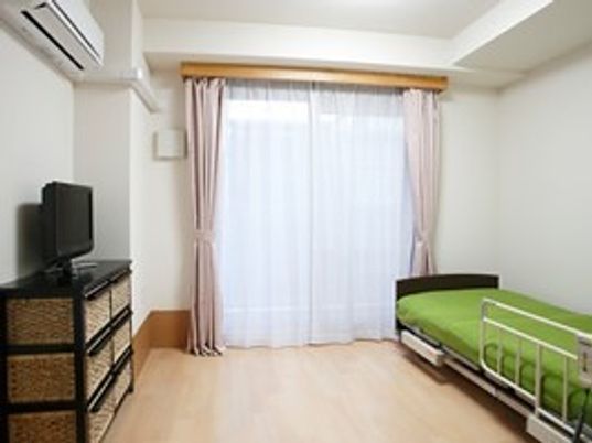 フローリングの居室。エアコンやベッド、テレビが設置され、大きな窓にはピンクのカーテンがある。