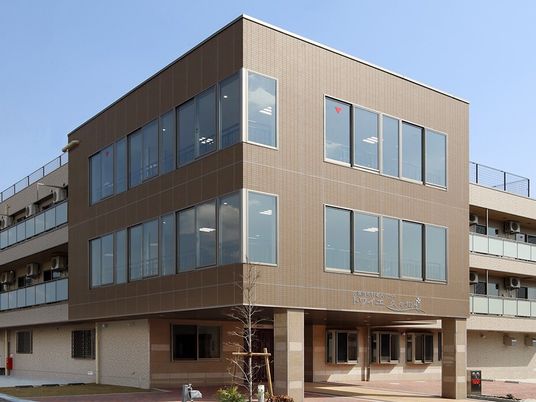 ３階建ての建物はブラウン系で各フロアにベランダがあり、建物の前には木が植えられている。