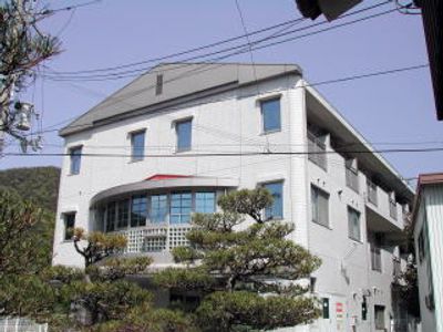 三角屋根が付いた、白を基調とした3階建ての建物。周辺には緑がたくさん植えられ、のどかな環境。