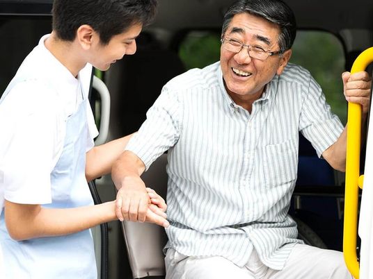 眼鏡をかけた高齢男性が福祉車両から下りようとしているところをサポートしている若いスタッフ。