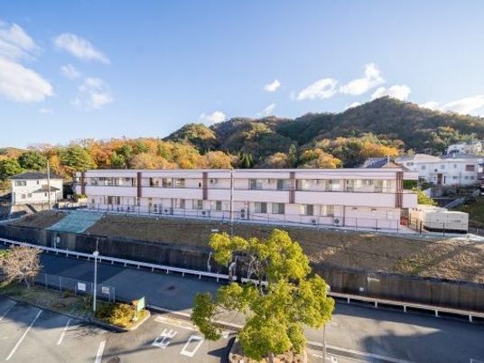 2階建ての建物は白を基調としていて、横に長い。周辺には山や自然があり、駐車場もある。