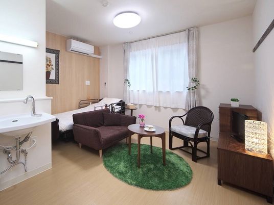 レースカーテンとカーテンが付いた大きな窓があるフローリングの部屋には介護ベッドや車いす対応の洗面所、タンスなどがある。