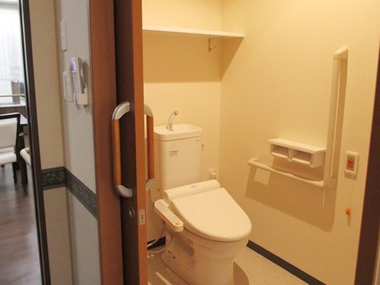 エル字型の手すりが付いたトイレ。オレンジの呼び出しボタンが付いた壁には二連のトイレットペーパーホルダーもある。