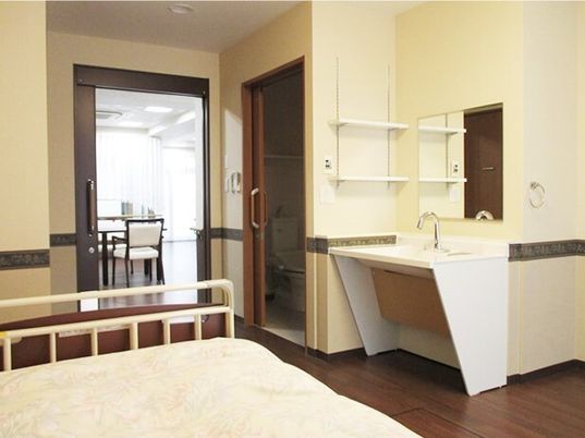 車いす対応の洗面所や介護ベッド、トイレがあるフローリングの部屋。スライド式のドアが付いている。