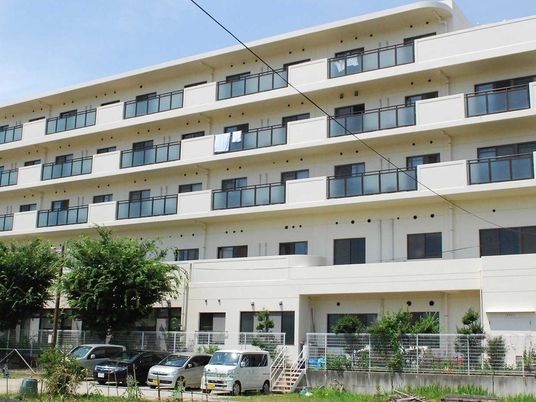 建物は白っぽい色で大きな病院のような雰囲気。建物の周辺には木が植えられ駐車スペースもある。