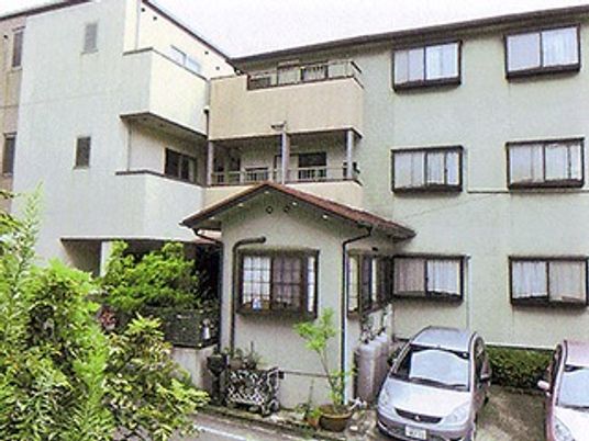 薄いグレーの3階建ての建物の各フロアにはベランダが付き、駐車スペースには数台の車が止まっている。