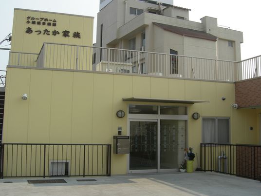 ホーム名と併設事業所名が書かれたベージュの外壁の建物は玄関に屋根やポストが付いている。