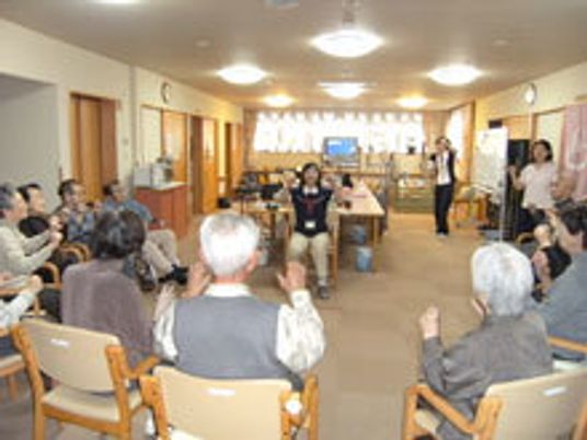 デイサービスでレクリエーションを楽しむ入居者とスタッフ。座ったまま体操をおこなっている。