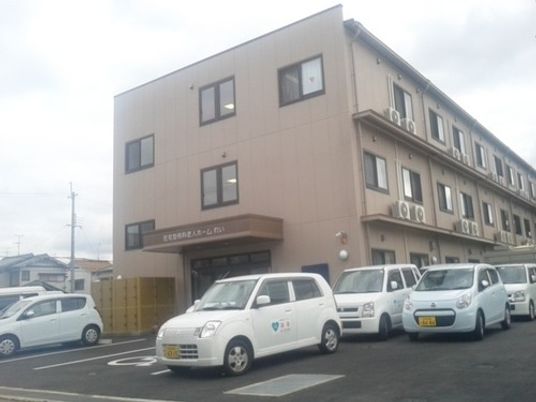 薄い茶色の外観をした建物。駐車場が広く、多数の自動車が停まっている。