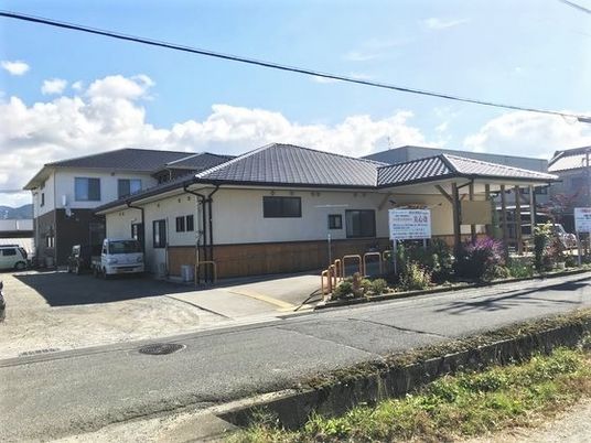 平屋建ての和風の建物の玄関前には屋根や木があり、奥には２階建ての建物がある。