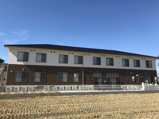 ２階建ての建物は白とブラウンの２色使い。周辺には田んぼが広がっている。