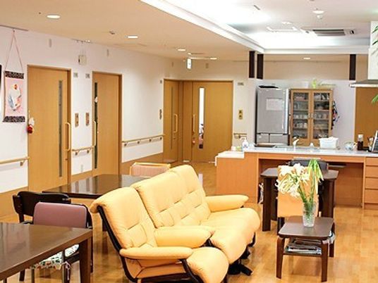 一軒家のリビングのような雰囲気の空間。大きなソファとテーブル、いすなどが置いてあり奥のほうにキッチンがある