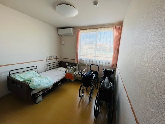 レースカーテンとカーテンが付いた窓がある部屋には寝具付きベッドやエアコン、車いすがあり、緊急通報装置付きで安全。