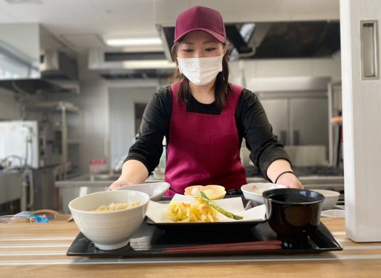 エンジのキャップとエプロンを付けた女性スタッフが天ぷらの盛り合わせや炊き込みご飯が盛られたトレーを差し出している。