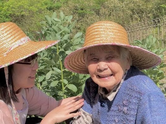 カメラ目線で麦わら帽子を来たおばあさんが笑っている。それを見つめるスタッフ