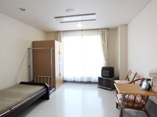 広めのワンルームタイプの部屋にベッドやタンス、テレビ、いすなどが置いてある様子