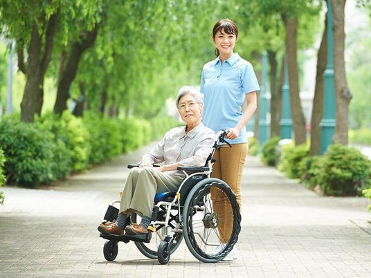 車いすの高齢女性と付き添いのポロシャツを着た女性スタッフ。遊歩道の中央に立ち止まり、微笑んでいる。