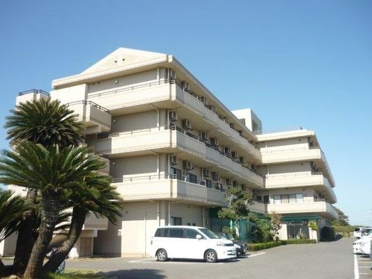 白っぽい建物は4階建ての頑丈な造り。敷地内には気が多く植えられ、車を止めるスペースもある。