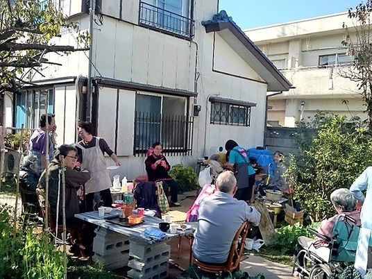 高齢者が庭に集まり、催し物を楽しんでいる様子。日差しが差し込んでいる様子が分かる