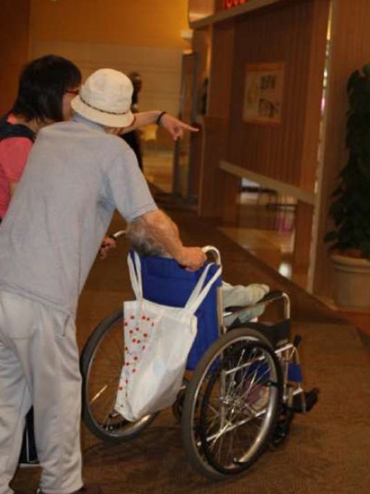 外出先で車いすに乗った入居者と、車いすを押しながら介助する帽子をかぶった男性。傍らにはスタッフがいる。