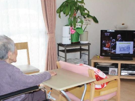ホームのリビングで国会中継のテレビを見ている女性入居者。テレビの脇には観葉植物が置かれている。