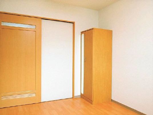 居室入口付近。角には縦長の収納家具、居室の出入り口は引き戸。壁は白、家具はライトブラウンに統一されている。