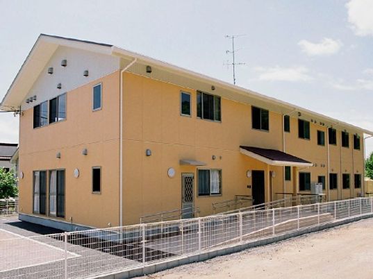 ２階建ての長方形の建物。三角の屋根と淡いオレンジ色の外壁が親しみやすい雰囲気。玄関周りもバリアフリー。