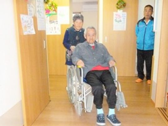 車いすに乗る高齢者とそれを押している家族。スタッフが見つめている様子