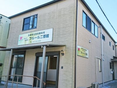 ブラウン系の外壁の2階建ての建物の玄関には手すり付きのスロープがあり、屋根も付いている。