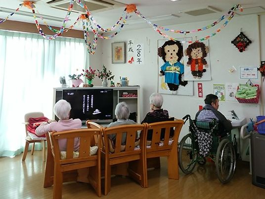 並んで木の椅子に座り、テレビを見ている様子の高齢者。車いすに乗る人、飾り付けられている室内