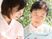 日当たりのいい場所でカルテのようなものを持って高齢女性と話をしているピンクのエプロンを付けた女性スタッフ。後ろには緑が生い茂っている。