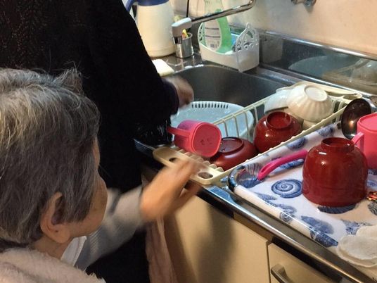 キッチンで皿洗いをしている人の隣で食器を並べる手伝いをしている車いすの入居者。