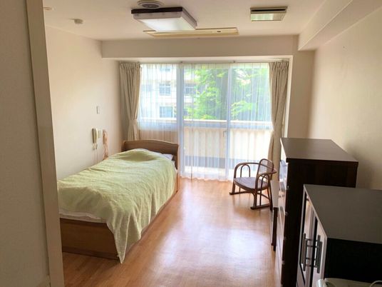 ベッドサイドには緊急通報装置が付いたフローリングの部屋にはタンスやいす、ベッドがあり、バルコニーも付いている。