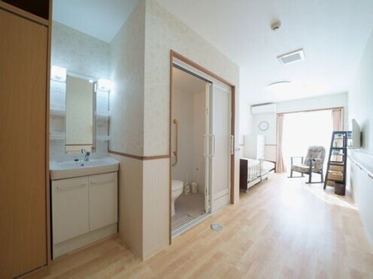 居室はフローリングでトイレや洗面所、クローゼットが付き、大きな窓のそばには寝具付きのベッドやいすもある。