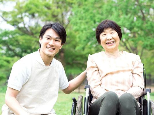 緑の多い屋外で笑顔で過ごしている車いすの高齢女性と付き添いの男性スタッフ。