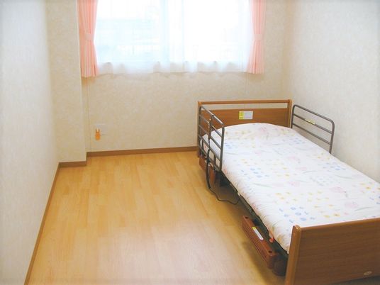 フローリングの居室。布団がセットされたベッドが置かれ、大きな窓にはピンクのカーテンがある。