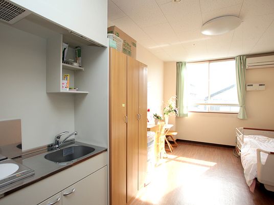 大きな窓があり明るい雰囲気のフローリングの居室。エアコンや寝具付きのベッド、クローゼットにミニキッチンが付いている。