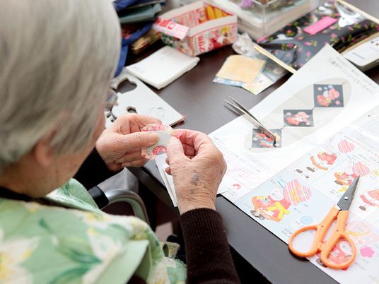 ハサミやピンセットを使用して細かい作業を真剣にしている高齢女性。見本のカラーコピーを熱心に見ながらしている。