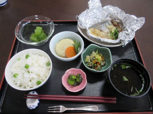 豆ごはんや魚のホイル焼き、煮物や漬物、すまし汁にフルーツとたくさんの小鉢が並んだ黒いトレーには、箸やフォークも置かれている。