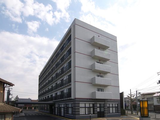 グレー系の大きな建物の各フロアにベランダがあり、エントランスには大きな車寄せが付いている。