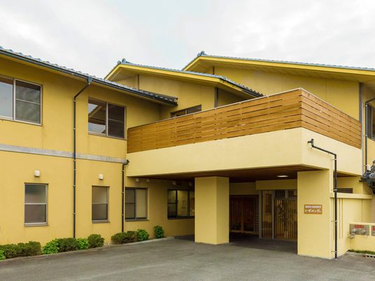 山吹色の外壁の２階建ての建物は昔ながらの日本家屋で建物の周りには低木が植えられている。