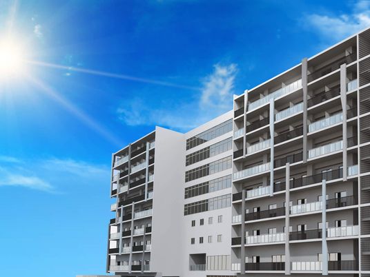 青空の下に建っている白を基調とした建物は９階建てで各フロアにベランダがあり、太陽の光が降り注いでいる。